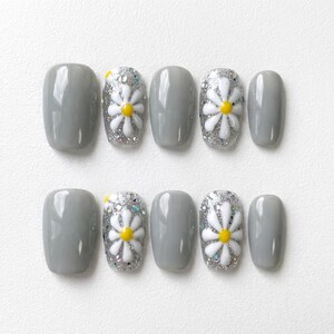 May include: A set of press-on nails in a light gray color. Some nails feature a white daisy design with a yellow center and silver glitter accents. The nails are oval-shaped and arranged on a white surface.