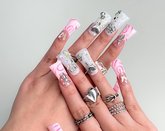 May include: A close-up of a hand with long, square-shaped acrylic nails. The nails feature a mix of designs, including pink swirls, clear glitter, and silver charms. Various silver rings adorn the fingers, adding to the overall aesthetic.