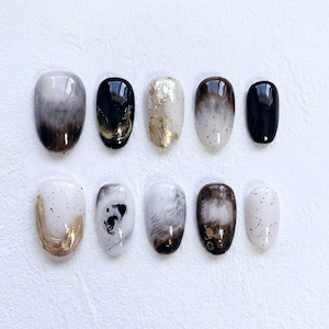 May include: A collection of ten artificial nails in various designs. The nails feature a range of colors, including black, white, gold, and brown. Some nails have a marbled effect, while others are solid or adorned with gold leaf or speckled patterns.
