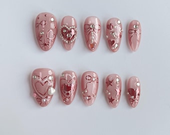 May include: A set of ten artificial nails in a light pink hue, adorned with heart and bow designs. Some nails feature silver rhinestones and pearl accents. The nails are oval-shaped and suitable for a manicure.