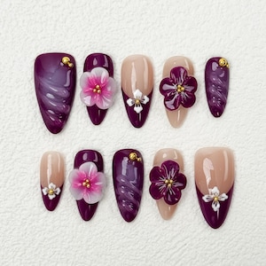 Peut inclure: Un ensemble de dix faux ongles de différentes conceptions. Certains sont d'un violet profond avec un effet marbré, d'autres sont nude avec des accents floraux blancs, et certains présentent des fleurs violettes en 3D avec des centres dorés. Chaque ongle est de forme ovale.