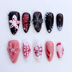 Könnte beinhalten: Eine Sammlung von zehn künstlichen Nägeln in verschiedenen Designs. Die Nägel weisen eine Reihe von Farben auf, darunter Schwarz, Rot, Weiß und Rosa, mit Verzierungen wie Perlen und floralen Mustern. Einige Nägel haben ein glänzendes Finish.