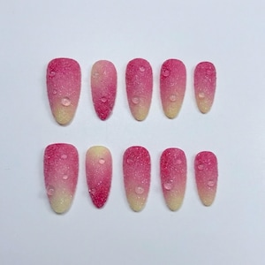Puede incluir: Un conjunto de diez uñas artificiales con un diseño degradado. Las uñas hacen la transición de un amarillo claro en la base a un rosa vibrante en la punta, con una textura brillante y un efecto de gota de agua.