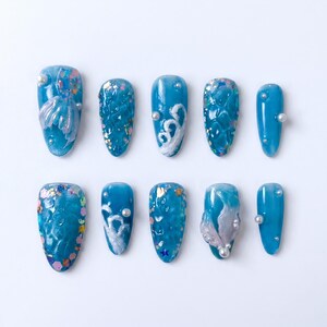 May include: A set of ten blue artificial nails with various designs. Some nails feature seashell and wave-like patterns, while others have glitter and pearl accents. The nails are arranged on a white surface.