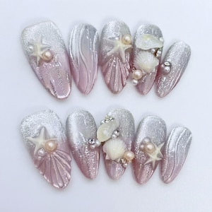 Könnte beinhalten: Ein Satz von zehn mandelförmigen Kunstnägeln. Die Nägel weisen ein Farbverlauf-Design aus silbernem Glitzer und Rosa auf, verziert mit Muschel- und Seestern-Verzierungen sowie Perlenakzenten. Die Gesamtästhetik erinnert an ein Strandthema.