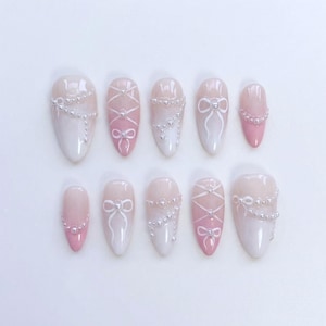 May include: A collection of ten artificial nails in varying sizes, with a pink and white gradient. Each nail is decorated with small silver bead details and white bow or criss-cross patterns, offering a delicate and elegant appearance.