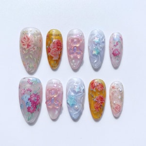 May include: A collection of ten oval-shaped artificial nails. Designs include floral patterns, teddy bear figures, and star accents. Colors range from pink and yellow to blue and clear, with pearl embellishments. The nails are arranged on a white surface.
