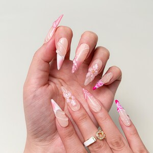 May include: A hand displaying long, almond-shaped acrylic nails. The nails feature a gradient design, transitioning from a nude base to a pink tip. Some nails have white swirls, pink polka dots, and 3D floral accents. A gold ring with a white band is visible.