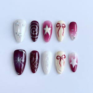 May include: A set of ten artificial nails in various designs. The nails feature a range of colors, including white, burgundy, and cream, with embellishments such as stars, bows, and swirls. Some nails have a gradient effect.