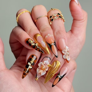 May include: A hand showcasing long, almond-shaped acrylic nails with diverse designs. The nails feature gold accents, floral details, and tortoiseshell patterns. Several gold rings are worn on the fingers, enhancing the overall aesthetic.