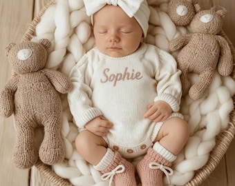 May include: A baby wearing a white onesie embroidered with the name "Sophie", a matching headband, and pink socks with white bows. The baby lies in a woven basket with knitted teddy bears and a chunky white knit blanket. The scene is soft and inviting.