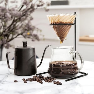 Peut inclure: Une cafetière à col de cygne noire, une carafe en verre avec du café, un système de café filtre et des grains de café sont disposés sur une surface blanche. La carafe a des mesures en onces liquides et en millilitres. Le système de café filtre comprend un support en métal noir et un filtre en papier.