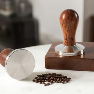 May include: Two coffee tampers with wooden handles and stainless steel bases. One tamper is upright on a wooden block, the other is lying on its side. A small pile of coffee beans sits nearby. The word "Daisy" is written on the handle of the upright tamper.