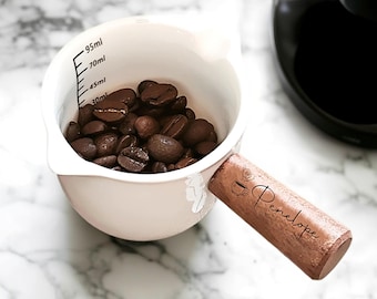 May include: A cream-colored measuring cup filled with coffee beans. The cup has a wooden handle with the name "Penelope" and a coffee cup graphic. The cup has measurement markings in milliliters.