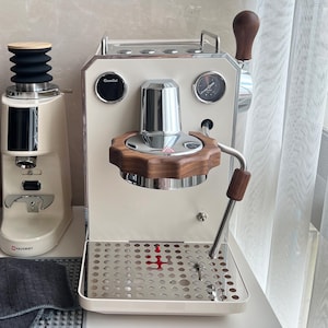 May include: Cream-coloured espresso machine with wooden handle and accents. A matching coffee grinder is to the left. The machine has a pressure gauge and a chrome portafilter. The drip tray is stainless steel.