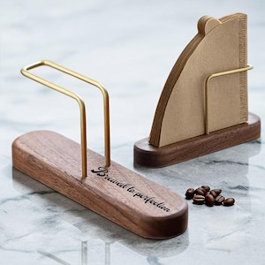 May include: Two wooden coffee accessories with gold-coloured metal accents. One holds coffee filters, the other has the text "Brewed to perfection." Coffee beans are scattered nearby. The items are on a marble surface.