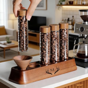 May include: A set of glass coffee bean storage tubes with wooden lids, displayed on a wooden holder. The holder has a built-in funnel and the initials "SL" with a deer antler design. One tube is being held up.