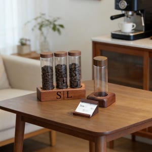 May include: A wooden coffee bean storage set with three glass tubes filled with coffee beans, a single glass tube, and a small card holder. The set is on a wooden table, with a coffee machine in the background.