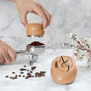 Könnte beinhalten: Ein Kaffee-Tamper aus Holz mit Edelstahlbasis wird über einem Siebträger mit Kaffeepulver gehalten. Der Tamper hat eine helle Holzoberfläche und ist mit den Buchstaben "A & J" und einem Blattmuster graviert. Kaffeebohnen sind verstreut.