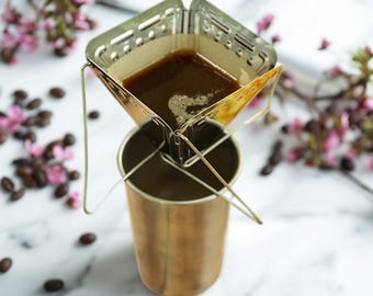 May include: A stainless steel pour-over coffee filter with a paper filter containing brewed coffee, positioned over a gold-coloured metal cup. The setup is on a marble surface, with coffee beans and pink flowers in the background.
