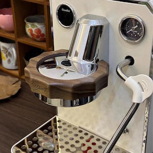 May include: A white and chrome espresso machine with a wooden base. The machine has a pressure gauge, a steam wand, and the name "Daisy" engraved on the wooden base. The machine also has the name "Gemilai" on the top.