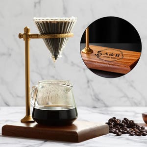 Peut inclure: Un support de cafetière à verser en métal doré avec une carafe en verre et un filtre conique en verre. La carafe est remplie de café noir. La base en bois porte les initiales "A & B" et la date "27.06.2023" gravées.