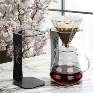 May include: A black metal pour-over coffee stand with the name "Michael" engraved on the side. A glass carafe with a handle and a clear glass coffee dripper are in use. The carafe contains brewed coffee.