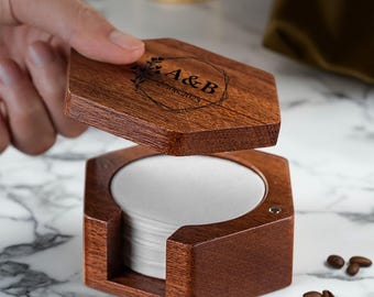 May include: A hexagonal wooden coffee filter holder with a hinged lid, engraved with the initials "A&B" and a date. Inside the holder is a stack of white paper filters. Coffee beans are scattered around the holder.