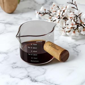May include: A clear glass measuring jug containing a dark liquid, with measurements in millilitres and fluid ounces. A wooden stopper with the word "Atticus" is placed on the side. White flowers are in the background.