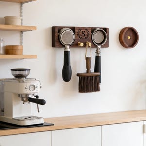 May include: A dark wood espresso station organiser with a monogrammed initial "S" and a laurel wreath. It holds two portafilters, a brush, and a tamper. A matching wooden lid and a cream-coloured espresso machine are also visible.