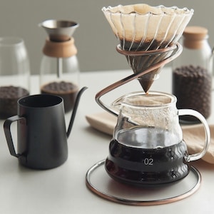 Peut inclure: Un ensemble de préparation de café avec une carafe en verre, un support en métal couleur cuivre et un cône filtre en papier. Une bouilloire noire et des bocaux en verre avec des grains de café sont également visibles. La carafe porte le numéro "02".
