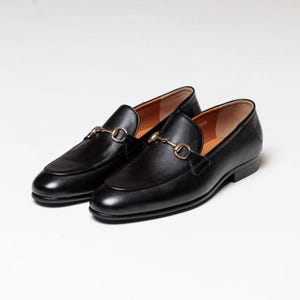 May include: A pair of black leather loafers with gold metal buckles. The loafers have a pointed toe and a low heel.