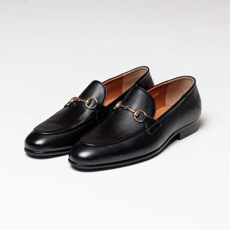 May include: A pair of black leather loafers with gold metal buckles. The loafers have a pointed toe and a low heel.