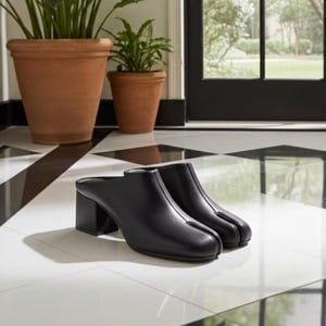 May include: A pair of black leather mules featuring a distinct split-toe design and a block heel. The shoes have a smooth finish, a closed-toe front with a vertical seam, and an open back, resting on a white and black tiled floor surface.