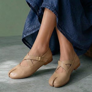 May include: Beige leather split-toe shoes featuring a textured finish, a low heel, and a cross-strap design across the top of the foot secured with a small gold-toned metal buckle. The footwear has a distinct cleft at the front toe area.