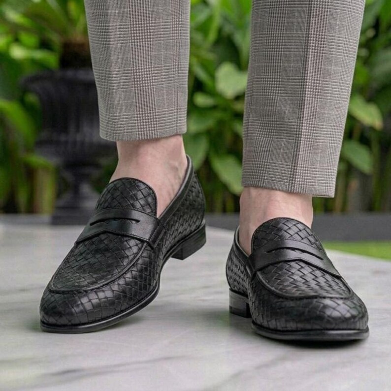 May include: Black woven leather loafers with a slip-on design. The shoes have a textured pattern and a low heel. The image shows the shoes from a low angle, with the wearer's legs in the background.
