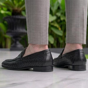 May include: Black leather loafers with a woven pattern. The shoes have a low heel and are worn with gray plaid trousers. The image is taken from a low angle, showing the shoes and the lower legs of a person.