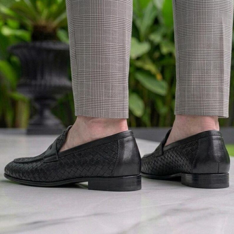 May include: Black leather loafers with a woven pattern. The shoes have a low heel and are worn with gray plaid trousers. The image is taken from a low angle, showing the shoes and the lower legs of a person.