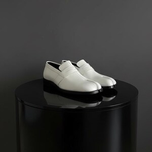 Pode incluir: Um par de mocassins de couro branco com sola preta e salto pequeno. Os sapatos são exibidos em um pedestal redondo preto sobre um fundo cinza escuro. Os mocassins têm um design clássico.