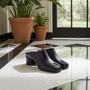 May include: A pair of black leather heeled mules with a split-toe design. The shoes have a block heel and are set on a black and white tiled floor. Two terracotta pots with green plants are in the background.
