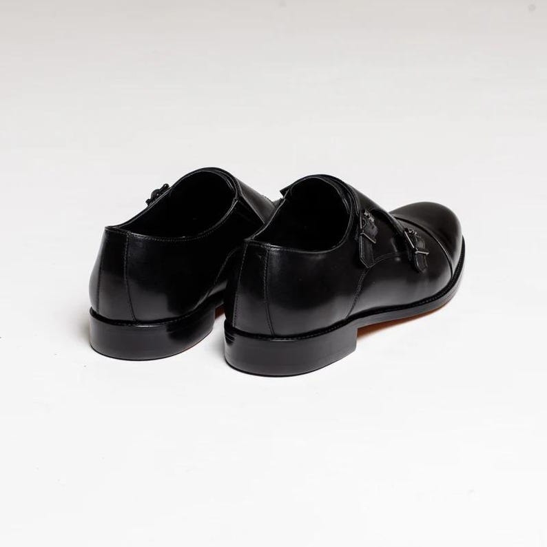 Puede incluir: Un par de zapatos de vestir de cuero negro con doble hebilla monje. Los zapatos tienen un acabado pulido y una punta cerrada.