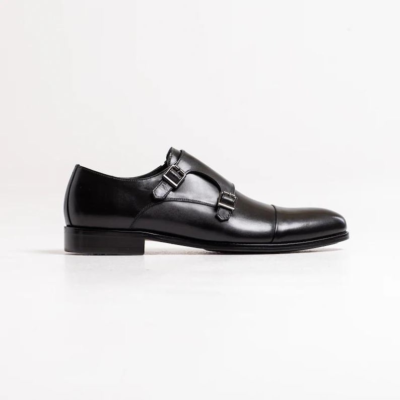 Puede incluir: Zapatos de vestir de cuero negro con doble hebilla monje y hebillas plateadas.