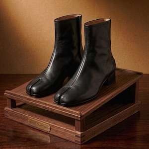 May include: A pair of black leather ankle boots with a distinct split-toe design, featuring a low block heel and a smooth finish. The boots are displayed on a rectangular wooden stand with a small metal plaque labeled MASTER CRAFTSMAN.