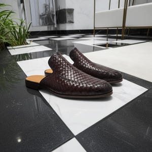 May include: A pair of dark brown woven leather slip-on shoes. The shoes have a low heel and a tan insole. The shoes are displayed on a black and white checkered floor. The shoes are a stylish footwear option.