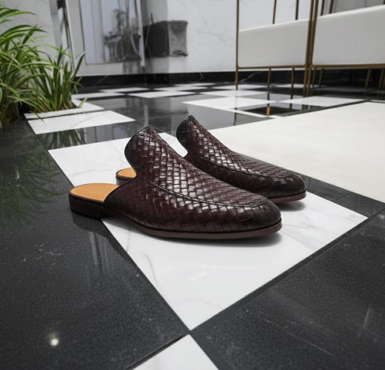 May include: A pair of dark brown woven leather slip-on shoes. The shoes have a low heel and a tan insole. The shoes are displayed on a black and white checkered floor. The shoes are a stylish footwear option.