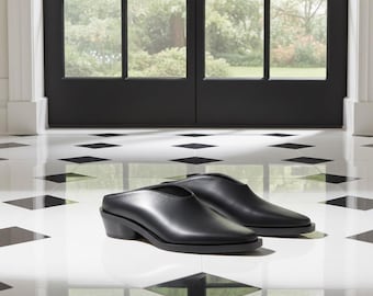 May include: A pair of black leather mules with a low heel. The shoes are positioned on a white and black tiled floor, with a black door and a view of greenery in the background.