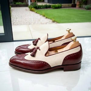 May include: A pair of two-tone leather loafers with a cream-colored suede upper and burgundy leather accents. The shoes feature decorative perforations, tassel details, and are displayed with wooden shoe trees.