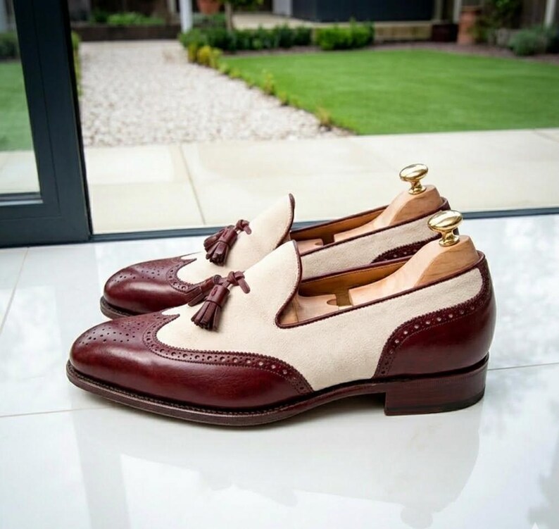 May include: A pair of two-tone leather loafers with a cream-colored suede upper and burgundy leather accents. The shoes feature decorative perforations, tassel details, and are displayed with wooden shoe trees.