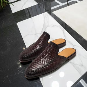 May include: A pair of burgundy woven leather slip-on shoes. The shoes have a rounded toe and a tan insole. The shoes are displayed on a black and white marble surface.
