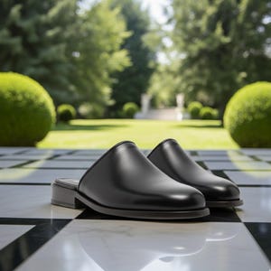 Op de afbeelding: Een paar zwarte leren muiltjes met een lage hak. De schoenen worden getoond op een zwart-wit geblokt oppervlak, met een vage groene achtergrond van bomen en verzorgde struiken.